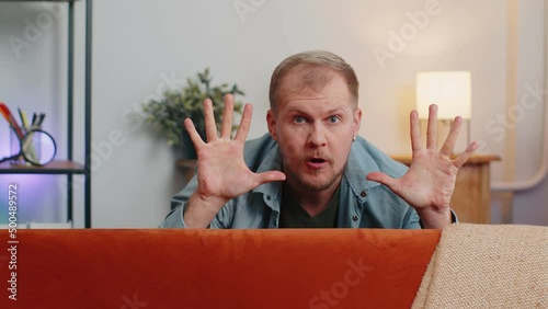 Portrait of cunning Caucasian man playing hide and seek peekaboo game hiding behind sofa looking at camera, smiling in positive mood. Fool around. Young adult guy play hide-and-seek game at home alone