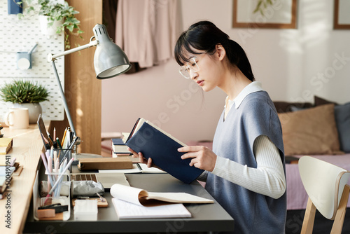 Concentrated Asian student girl in eyeglasses and knitted vest sitting at table and reading textbook while preparing for exam