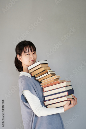 Portrait of puzzled Asian student girl in knitted vest carrying big stack of books against isolated background