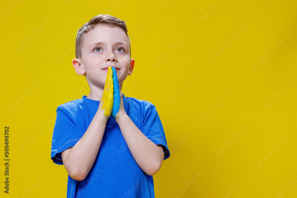 Ukrainian boy prays for Ukraine. Children against war. A boy in a blue T-shirt on a yellow background folded his hands painted in the Ukrainian flag for prayer Ukrainian boy prays for Ukraine. Children against war. A boy in a blue T-shirt on a yellow background folded his hands painted in the Ukrainian flag for prayer