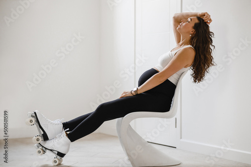 Healthy pregnant European woman staying near white wall on roller skates. Pregnant lady with white top and tight black pants. Relaxing exercise. Cheerful expecting mother. Healthy lifestyle.