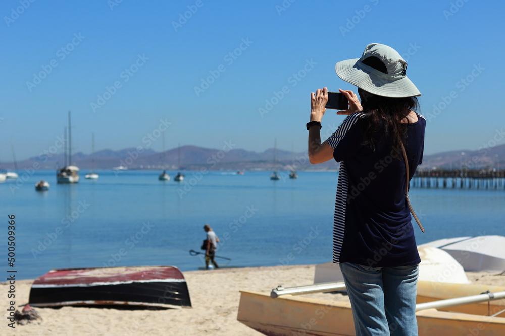 A young woman by the sea takes a photo of sailboats anchored in the bay. A fisherman at the water's edge