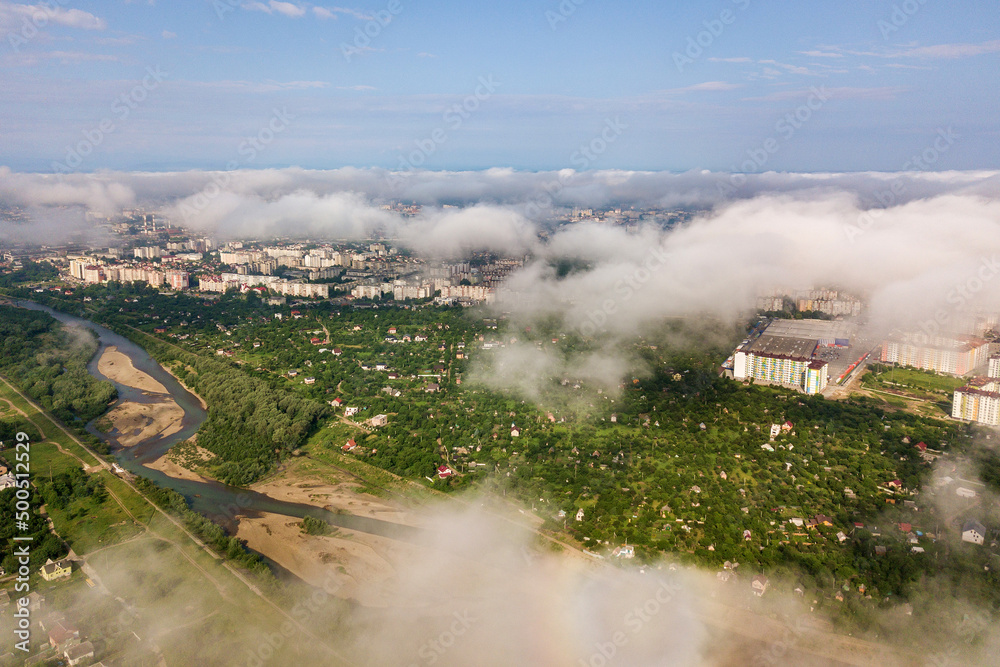 Fototapeta premium Aerial view of white clouds above a town or village with rows of buildings and curvy streets between green fields in summer. Countryside landscape from above.