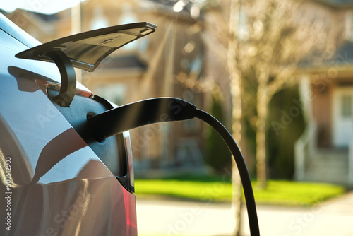 EV electric car plugged charging at residential home under sunset light against blurred street and house around background. Close up.