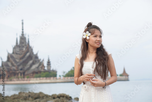 Portrait of beautiful Asian woman in white dress smiling on the beach with the frangipani flower. Asian stylish happy woman relaxing at tropical beach during summer vacation. travel concept. photo