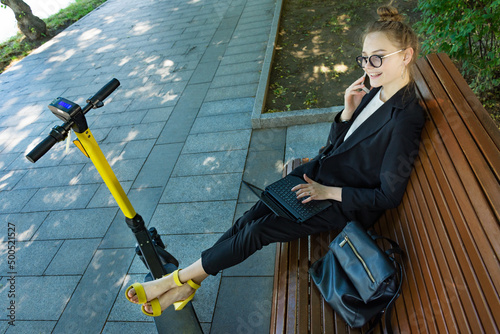 Businesswoman in a black business suit sits on a park bench with a laptop and a phone, working, putting her feet on her electric scooter on which she arrived. Electric scooter. Alternative transport.