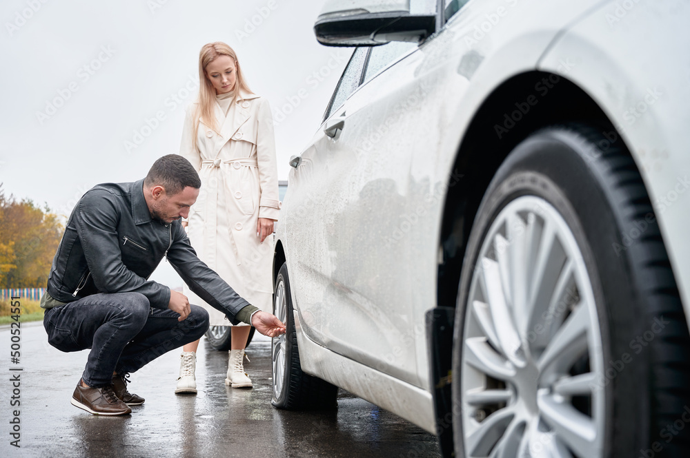 Young man unscrewing cap from nipple of rear lowered wheel, blonde woman standing next to him and looking at damaged tire. Preparing wheel for pumping and restoring internal pressure. Young man unscrewing cap from nipple of rear lowered wheel, blonde woman standing next to him and looking at damaged tire. Preparing wheel for pumping and restoring internal pressure.