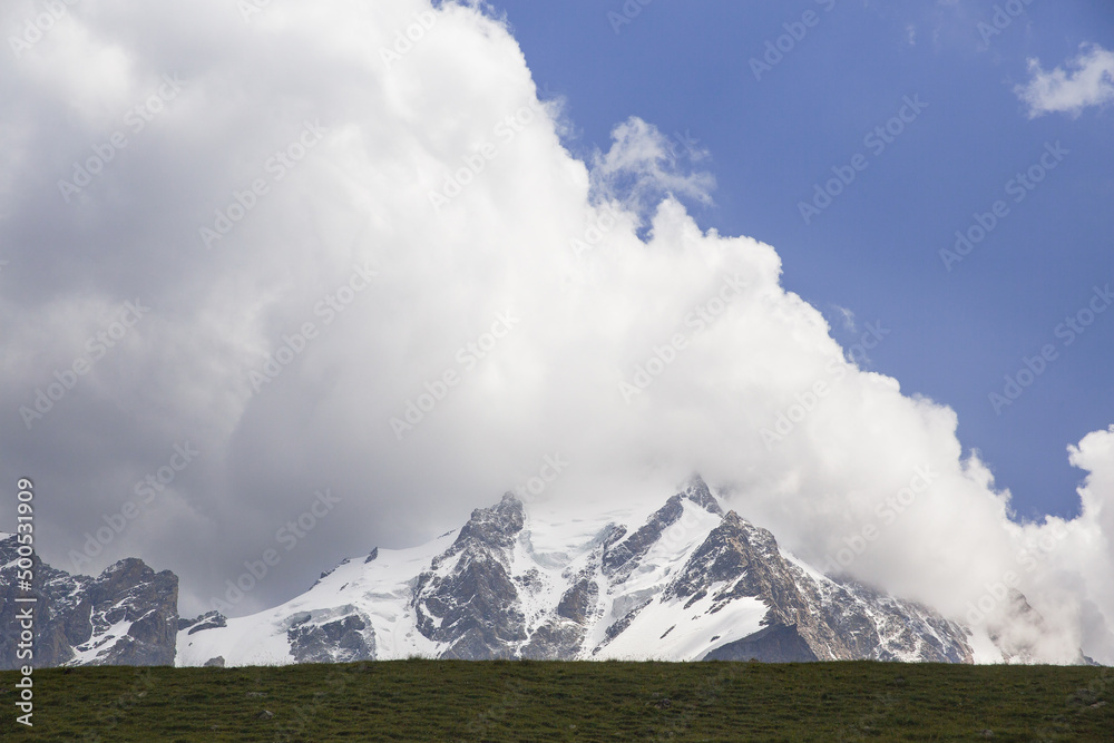 Snowy peak of the mountain against the background of the sky with clouds. At the bottom of the frame is a narrow strip of land.