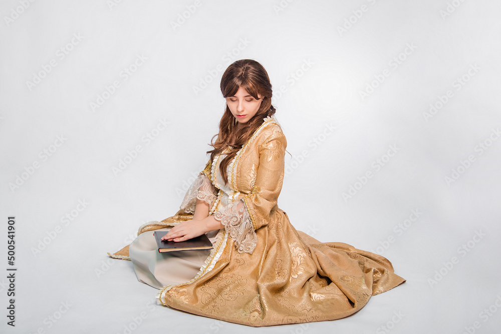 Naklejka premium Full length portrait of a young woman in a puffy gold dress in the rococo era, posing while sitting with a book in her hands, isolated on a white background. The girl is reading a book.