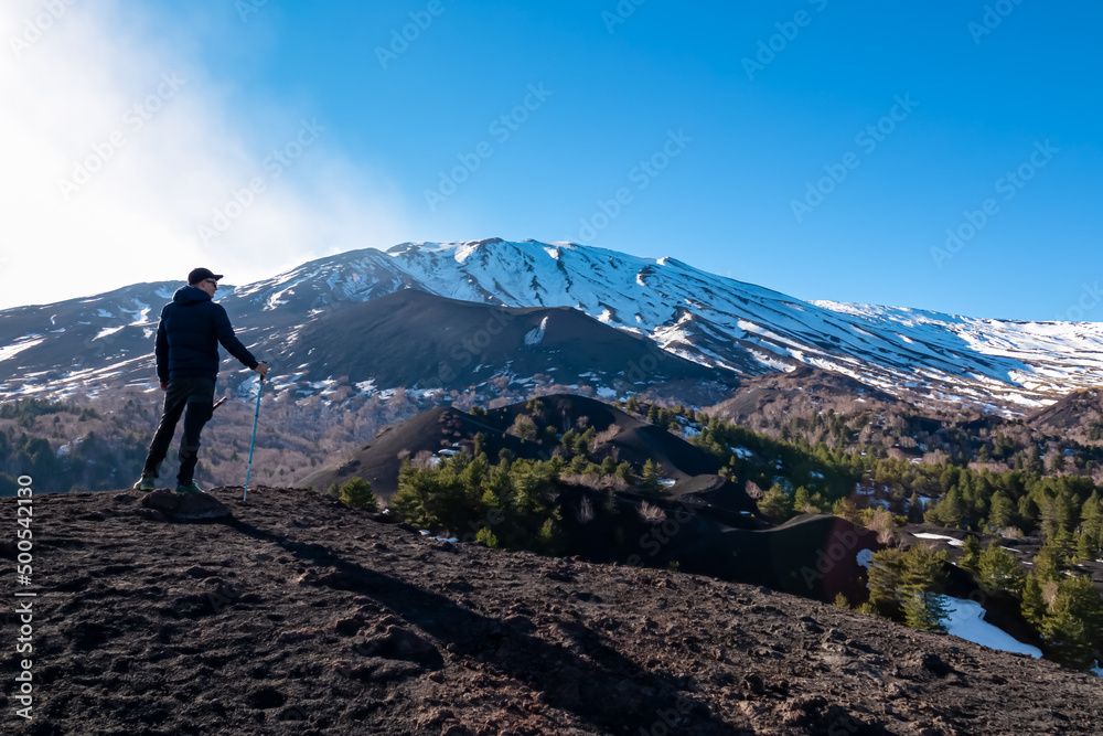 Man with hiking pole on volcanic landscape of volcano mount Etna, in Sicily, Italy, Europe. Pine and white birch trees growing on solidified lava, ash and pumice. Slopes of crater covered with snow