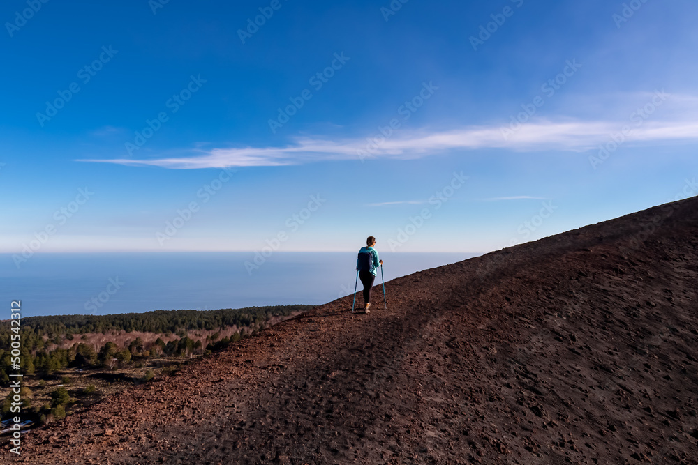 Tourist woman hiking around the crater of Sartorio on volcanic landscape of volcano mount Etna, in Sicily, Italy, Europe. View on the Ionian sea. Walking on solidified lava, ash and pumice ground