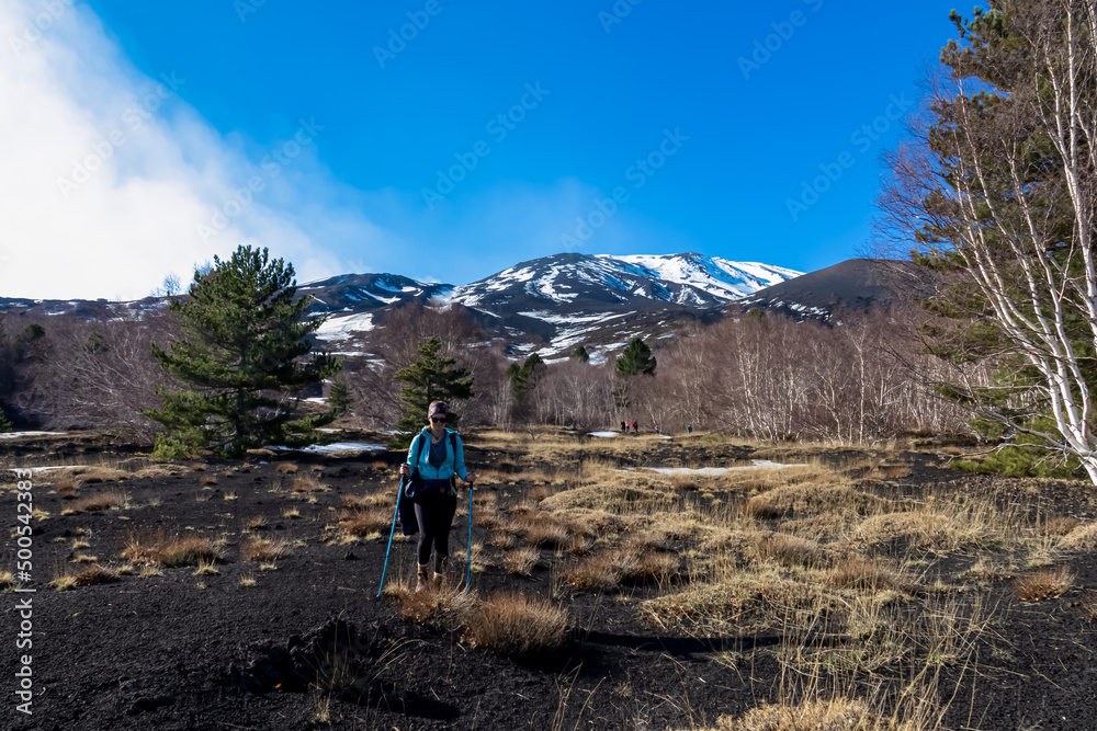 Tourist woman hiking on volcanic sand of volcano mount Etna, in Sicily, Italy, Europe. Yellow golden dry grass on bare terrain,  solidified lava, ash and pumice. Slopes of crater are covered with snow