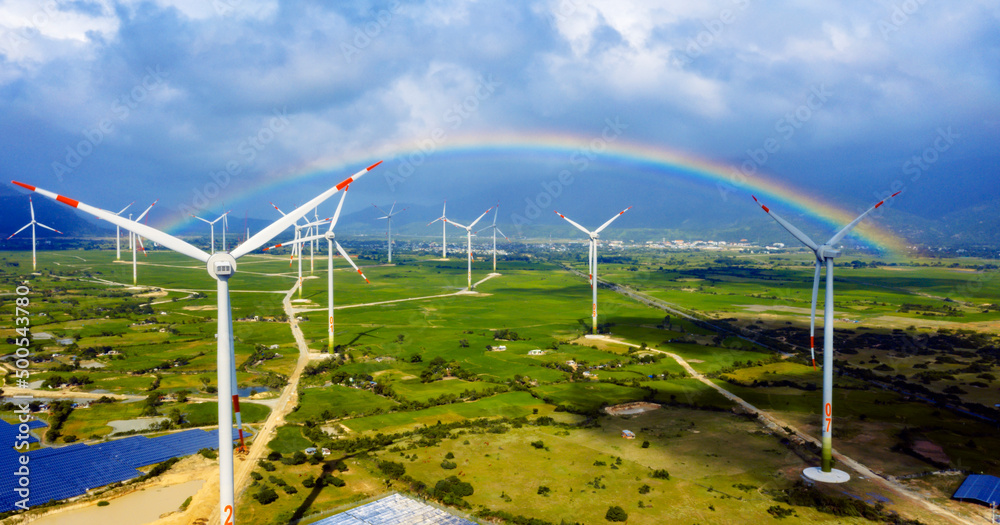 Wind turbine towers in a green field under the blue sky with rainbow generating alternative energy for sustainable growth. Reducing climate change and global warming Wind turbine towers in a green field under the blue sky with rainbow generating alternative energy for sustainable growth. Reducing climate change and global warming