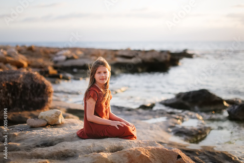 A European teenage girl in a dress sits on stones on the seashore, peace and quiet, a lonely child looks at the sea