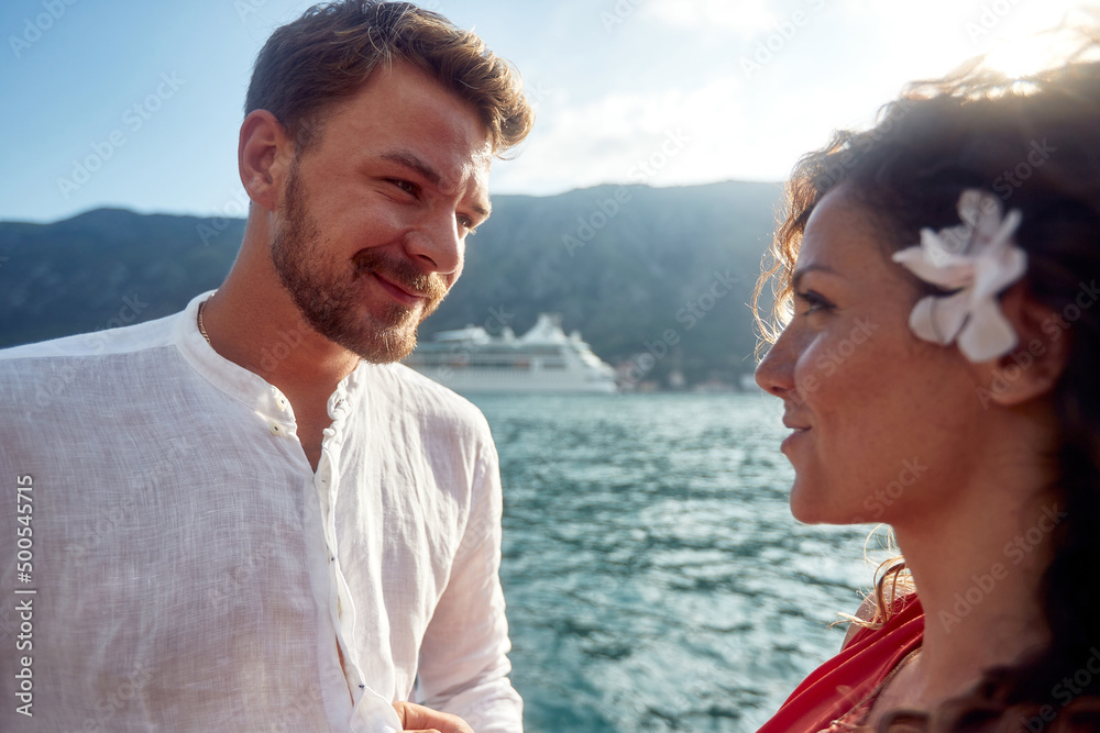 Portrait shot of young couple looking at each other. Cruiser and mountains in background. Summertime holiday by seaside. Togetherness, lifestyle, love, holiday concept.