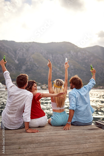 Two young couples having a good time with the drink while sitting on the dock on the seaside. Friends, friendship, holiday, sea