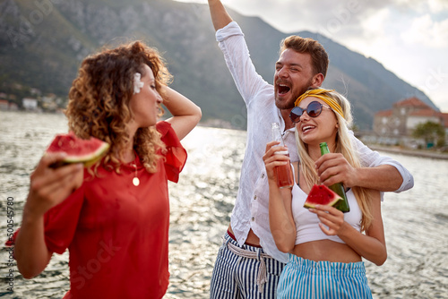 A group of friends having a good time while enjoying drink and juicy watermelon on the dock on the seaside. Friendship, holiday, sea © luckybusiness