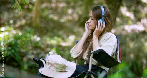 Young asian woman sit in a chair near the stream, listening to music from tablet with wireless headphones happily while camping in the woods