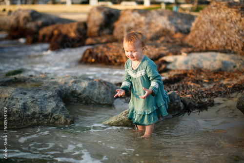 Fototapeta Naklejka Na Ścianę i Meble -  Toddler girl in a dress plays with water on the seashore at sunset, sand and stones, lifestyle