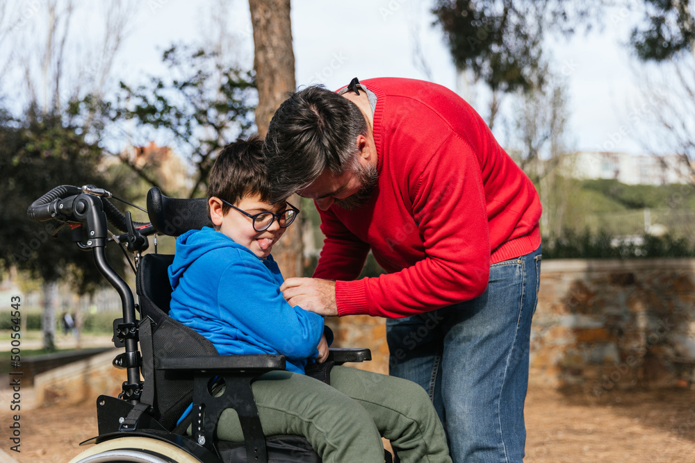 Father playing with his son with disability in a wheelchair while they enjoy time together outdoors in the park. Father playing with his son with disability in a wheelchair while they enjoy time together outdoors in the park.