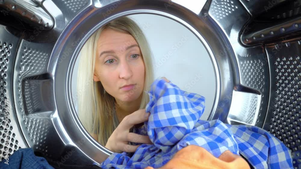 Young Caucasian housewife looks into drum of washing machine, takes out shirt with hole on it. Washing machine ruined clothes, tore them up. Woman is very surprised and upset. Call wizard, warranty Young Caucasian housewife looks into drum of washing machine, takes out shirt with hole on it. Washing machine ruined clothes, tore them up. Woman is very surprised and upset. Call wizard, warranty