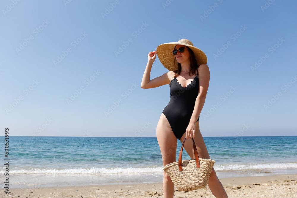 Young beautiful woman wearing one piece swimsuit, broad brim straw hat posing with a beach bag. Female fashion model at sandy beach on beautiful sunny day. Mediterranean sea background. Copy space Young beautiful woman wearing one piece swimsuit, broad brim straw hat posing with a beach bag. Female fashion model at sandy beach on beautiful sunny day. Mediterranean sea background. Copy space