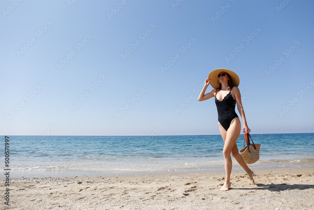 Young beautiful woman wearing one piece swimsuit, broad brim straw hat posing with a beach bag. Female fashion model at sandy beach on beautiful sunny day. Mediterranean sea background. Copy space Young beautiful woman wearing one piece swimsuit, broad brim straw hat posing with a beach bag. Female fashion model at sandy beach on beautiful sunny day. Mediterranean sea background. Copy space