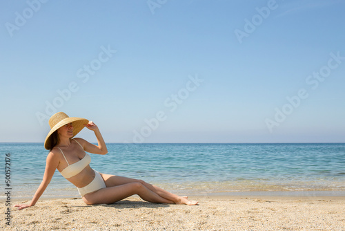 Caucasian woman with fit body in a bikini bathing suit with high waisted bottom and broad brim straw hat posing at sandy beach on beautiful sunny day. Mediterranean sea background. Copy space for text