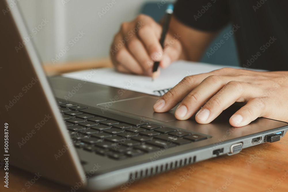 A businessman calculating money, money planning, loan budget, online payment concept on a laptop and take notes in a notebook with a pencil, working paperwork at the desk. Work from home.