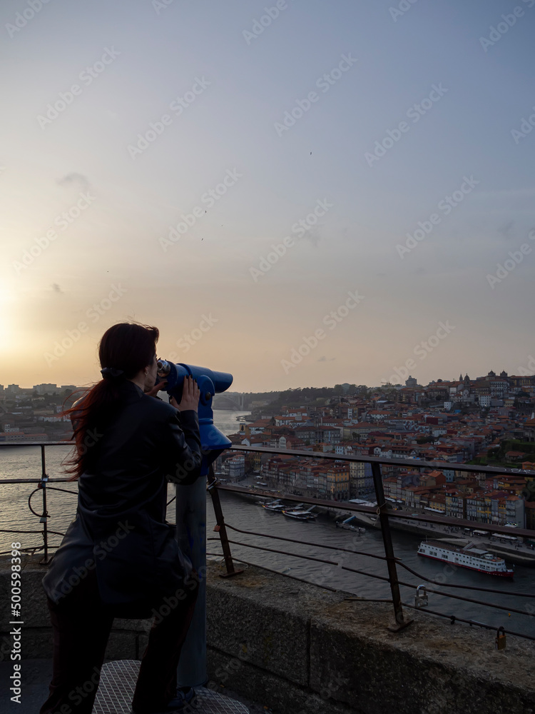 Fototapeta premium Young woman looks through coin binoculars to see Porto town in Portugal with Douro river. Traveler enjoying view panoramic point on vacation. Tourist at sunset visiting a scenic destination and using.