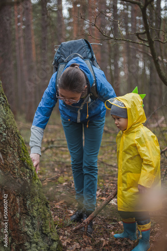 Mom and child walking in the forest after rain in raincoats with wooden sticks in hands