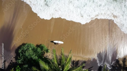 4K drone view from above surfer walking along sandy beach with surfboard and waves rushing in. Sports concept, vacation, spending time outside.