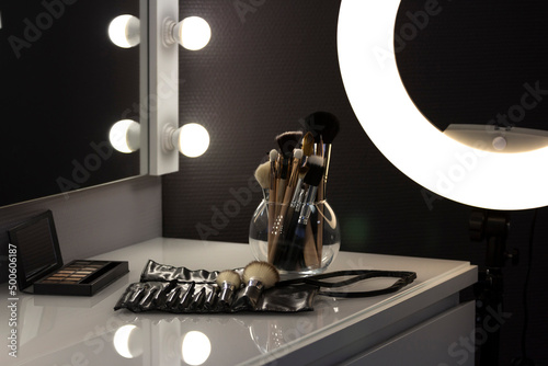 Mirror with light bulbs and ring lamp, near cosmetic brushes on dressing table indoors in makeup room. Shooting in a low key