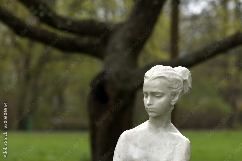 White marble sculpture face of young woman. Tree and spring, green grass in background. White marble sculpture face of young woman. Tree and spring, green grass in background.