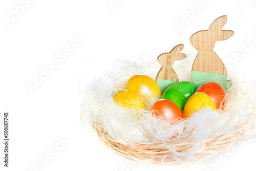Сomposition for easter in a wicker basket colorful eggs and wooden bunnys on white background. Copy space.