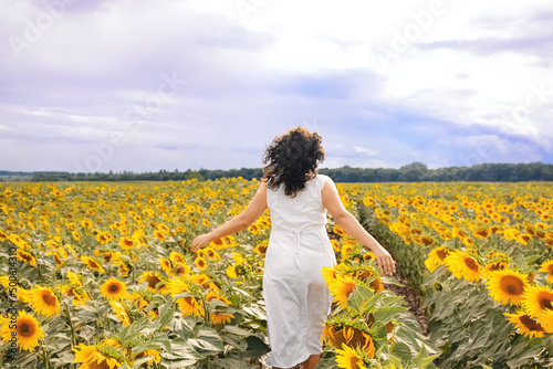 Back view Beautiful girl with curly hair in a white dress walks running through a field of yellow blooming sunflowers. Blue gloomy sky before the storm. Soft selective focus on photo.