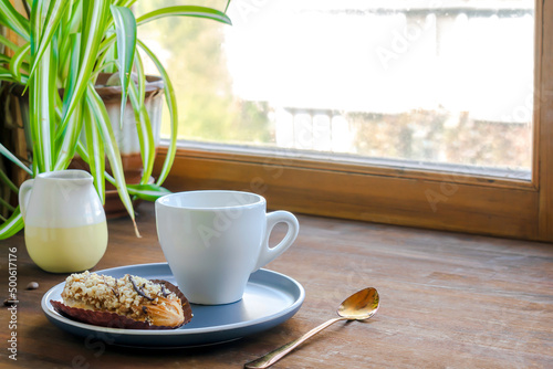 white espresso coffee cup  fresh baked eclair sweet dessert on plate kitchen table against window  utensils dishware  milk jug  home green plant. morning french home breakfast  copy space