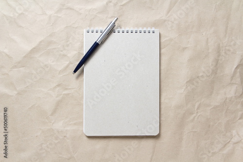 Gray notepad with white coiled spring and pen on a background of beige crumpled craft paper. With empty space for text and design