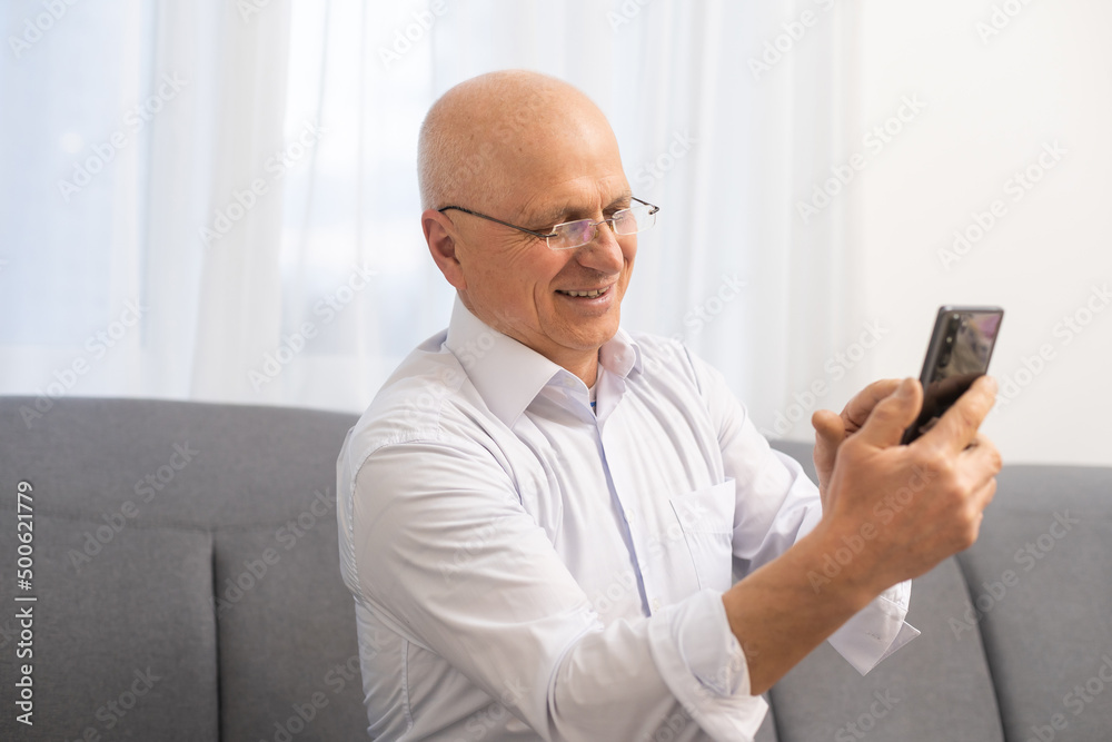 Handsome old man dressed in smart casual style and eyeglasses is using a smartphone and smiling while sitting on couch at home. Handsome old man dressed in smart casual style and eyeglasses is using a smartphone and smiling while sitting on couch at home.