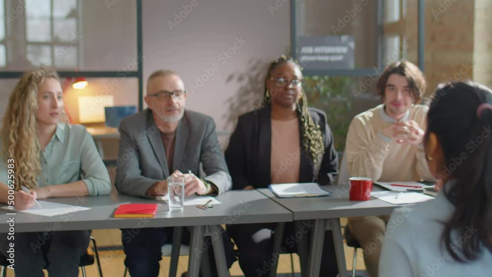 Rack focus shot of female job candidate sitting in front of multiethnic recruitment committee and giving them self-presentation during interview in office