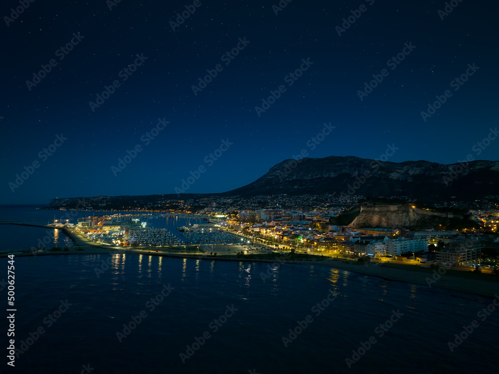 Denia on the Costa Blanca in the evening after the blue hour. A drone shot from the sea overlooking the harbour.
