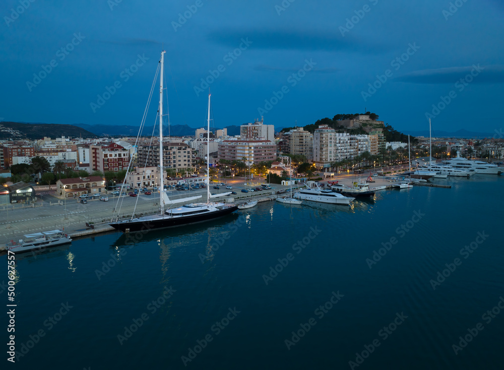 Denia on the Costa Blanca early in the morning at the blue hour. Ships are in port. In the foreground is a large sailing yacht.