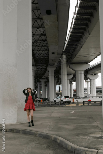 a girl in glasses, a black leather jacket and a red dress is standing under the overpass