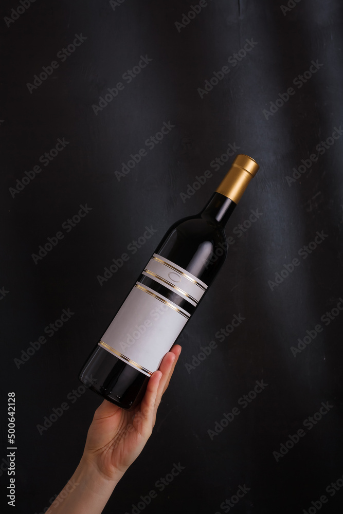 Fototapeta premium hand holding bottle of red wine with mock up label, no brand on black background. Cabernet sauvignon, merlot, pinot noir. Alcohol drink, copy space