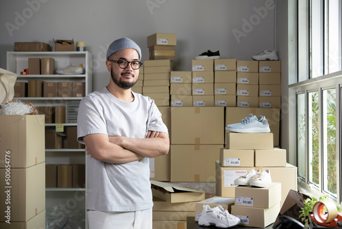 Confident young Asian man retail seller, entrepreneur, online store drop shipping small business owner looking at camera standing in delivery shipping warehouse with parcel boxes. photo