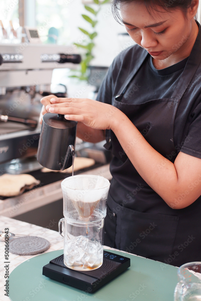 Smiling Asian barista young woman is wearing apron and pouring and craft a hot black coffee into cup for according to the customer's order at counter bar in coffee shop.
