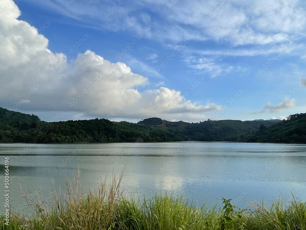 Perfect landscape. Amazing panoramic view. Lake, green grass, hills, covered by forest, blue sky with beautiful clouds, reflection of skies on the calm water surface. Nature at summer. Countryside.  