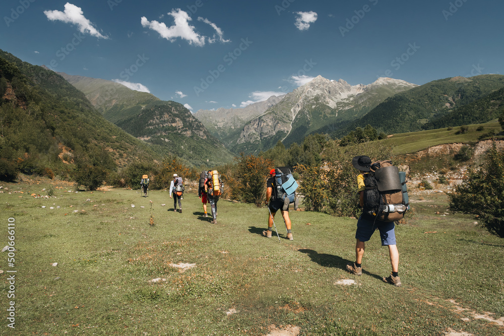 A group of tourists with large backpacks climbs the grassy slope overlooking the snow-capped peaks in the Caucasus Mountains Georgia Svaneti