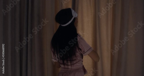 Woman with long loose hair in satin pink pajamas is getting ready to sleep in the bedroom.Brunette lies down on the bed, closes the curtains, covers herself with a white blanket and puts on sleep mask