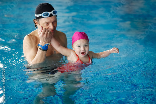 Happy kid toddler learns to swim with coach in the pool, dad grandpa teaches to swim daughter in pink swimming cap